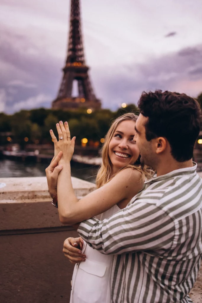Eiffel Tower engagement photoshoot of a couple at dusk, the woman showing her new ring as she smiles at her fiancé with the glowing tower and the Seine behind them