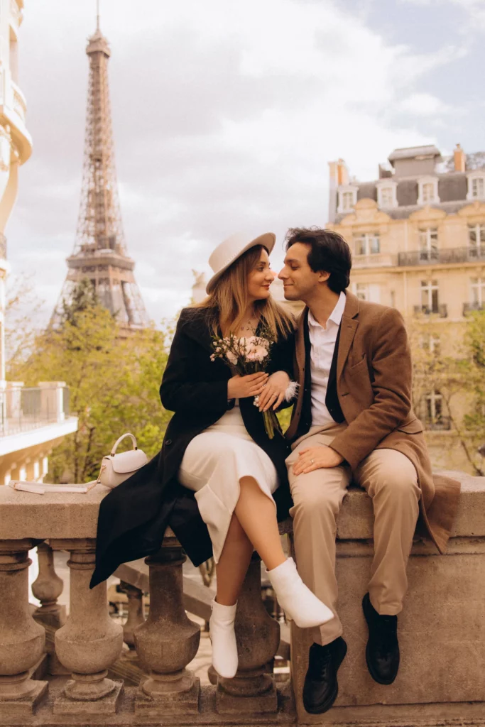 Spring couple photoshoot in Paris, a couple leaning in for an almost kiss on a stone balustrade with the Eiffel Tower soft behind them and fresh green trees around