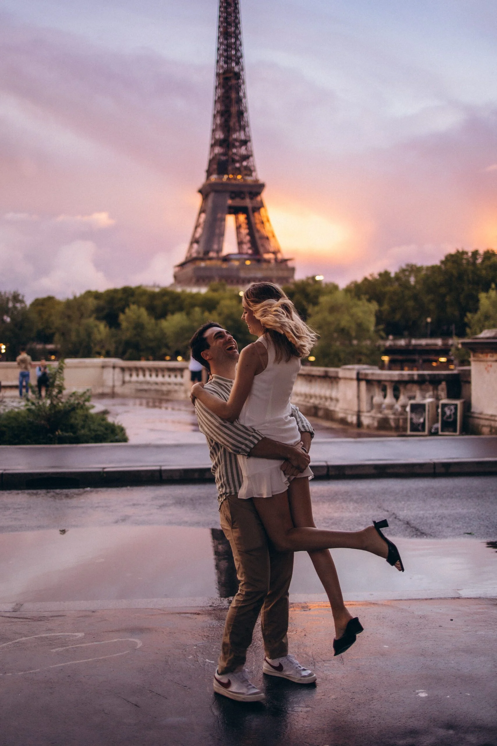Sunset Eiffel Tower photoshoot of a couple, the man lifting his laughing partner on the wet stone of Trocadéro as the sky glows pink behind the tower