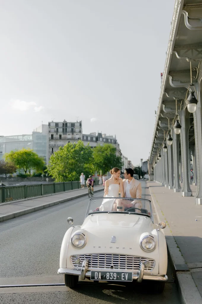 Paris couple photoshoot in a vintage white Triumph convertible on the Bir-Hakeim bridge, a couple in a pale gown and beige suit, by Julia Litvin