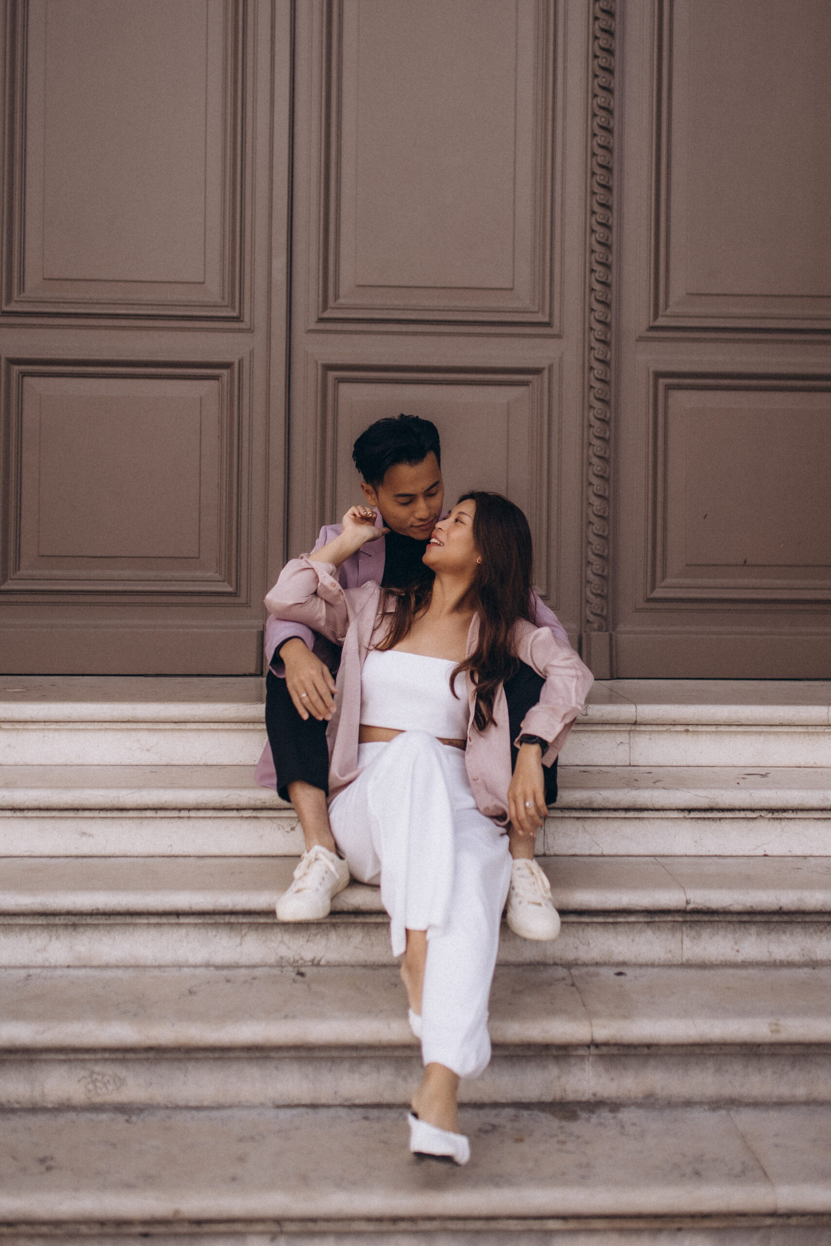 Paris engagement photoshoot, a couple sitting close on grand marble steps, the man leaning to her cheek as she smiles up at him, her engagement ring catching the light, in a soft pink and white palette