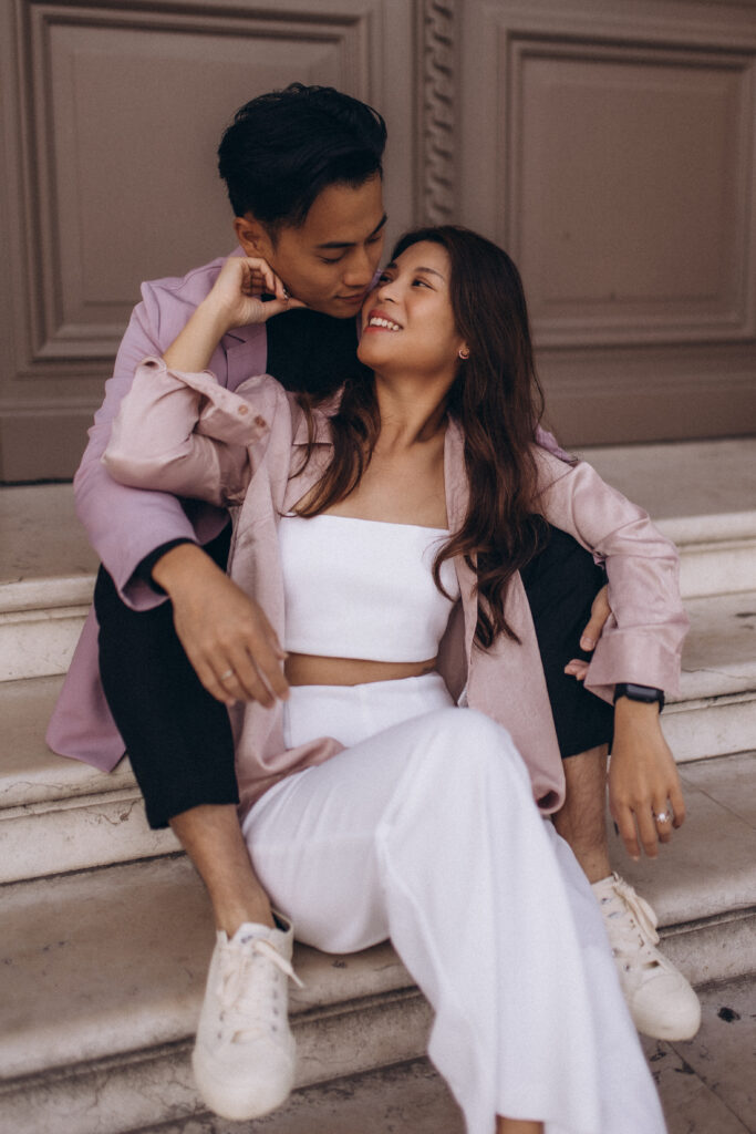 A couple at their Paris engagement session sitting on stone steps and gazing at each other, the woman in white laughing softly, an engagement ring on her hand