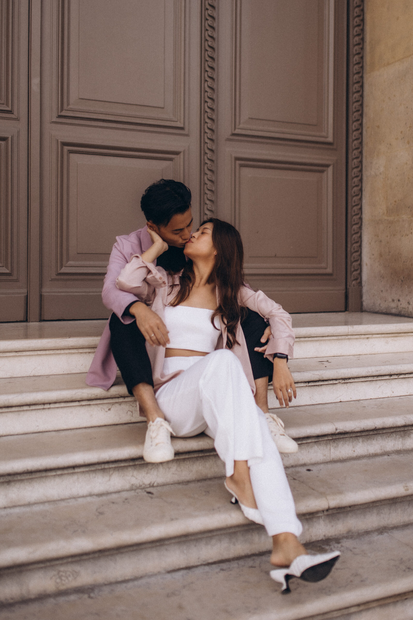 An engagement photoshoot in Paris, a couple kissing on marble steps before tall paneled doors, the woman's white heel slipping free, in warm neutral tones