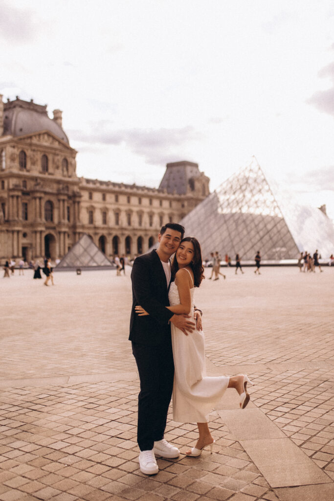 Louvre couple photoshoot in Paris, a couple embracing and laughing in front of the Louvre palace and its glass pyramid, the woman lifting one heel