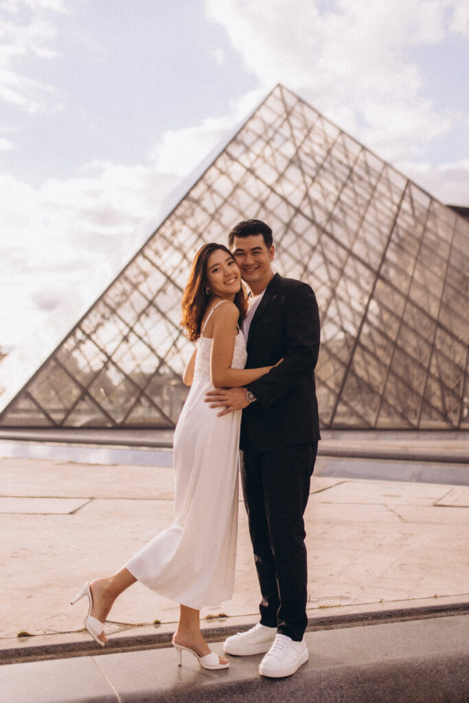 Paris couple photoshoot at the Louvre, a couple in a white dress and black suit embracing in golden light before the glass pyramid