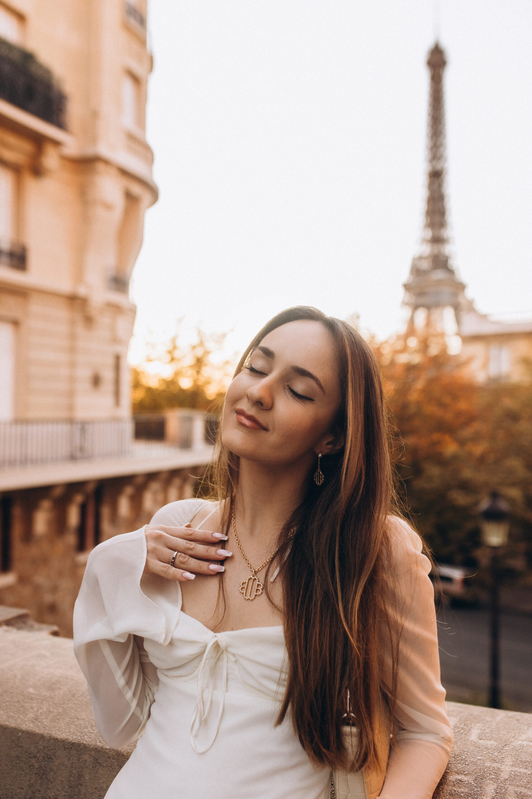 A woman in a white dress at golden hour, eyes closed, by a stone balustrade with the Eiffel Tower blurred behind her.