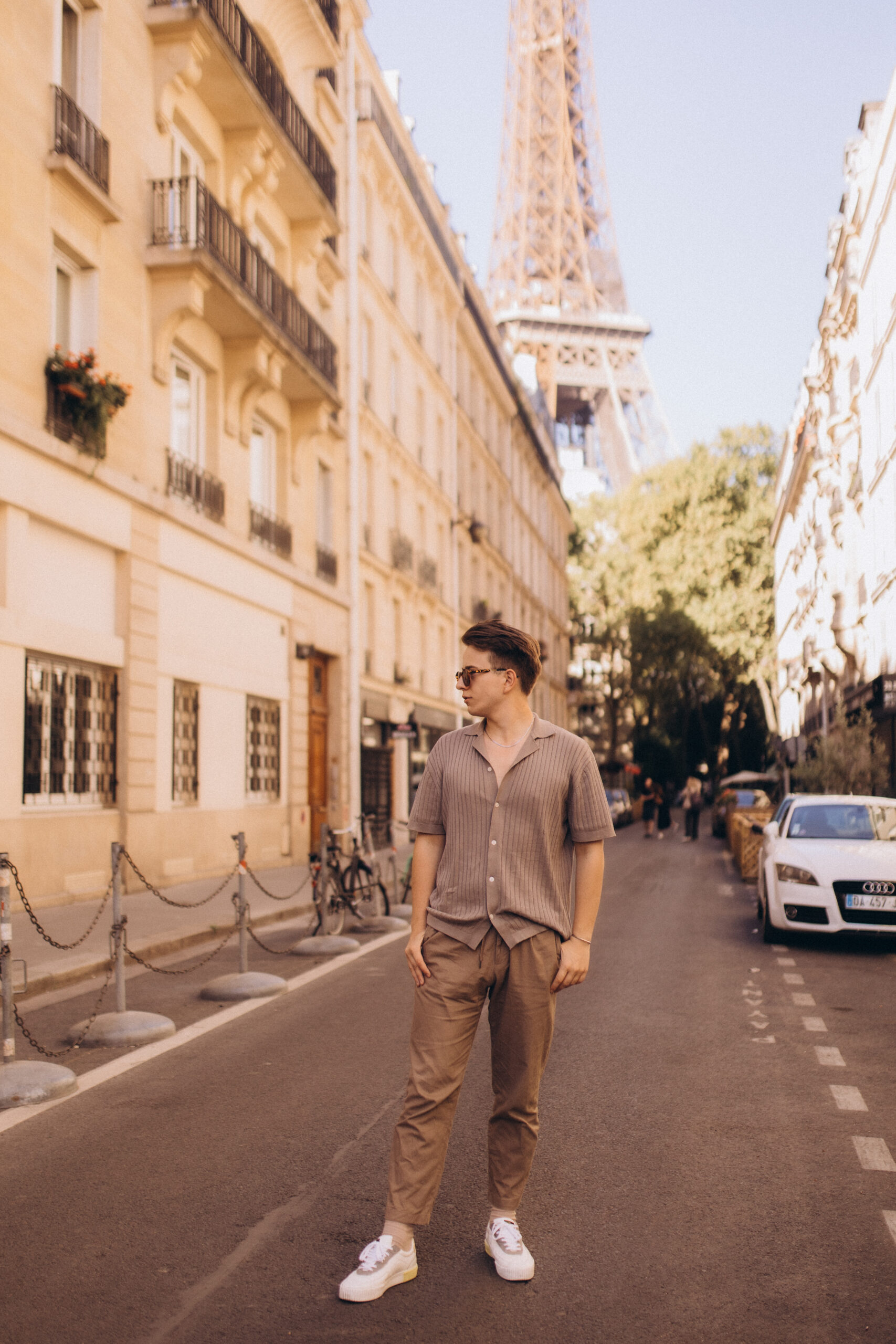 Paris street photography, a young man in a ribbed shirt and beige trousers standing in the middle of a quiet Parisian street with the iron tower rising at the far end