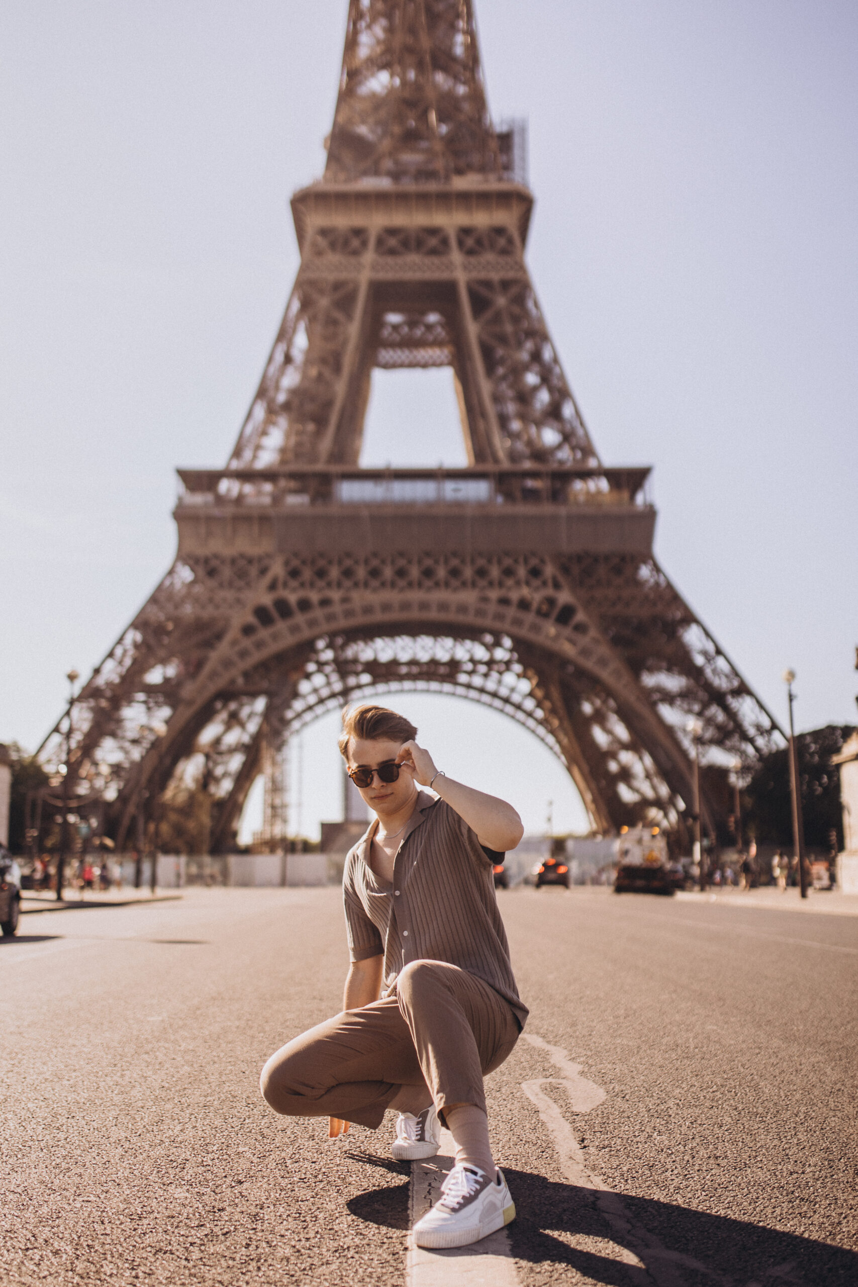 A young man crouching on an open esplanade directly beneath the Eiffel Tower, adjusting his sunglasses in bright daylight.