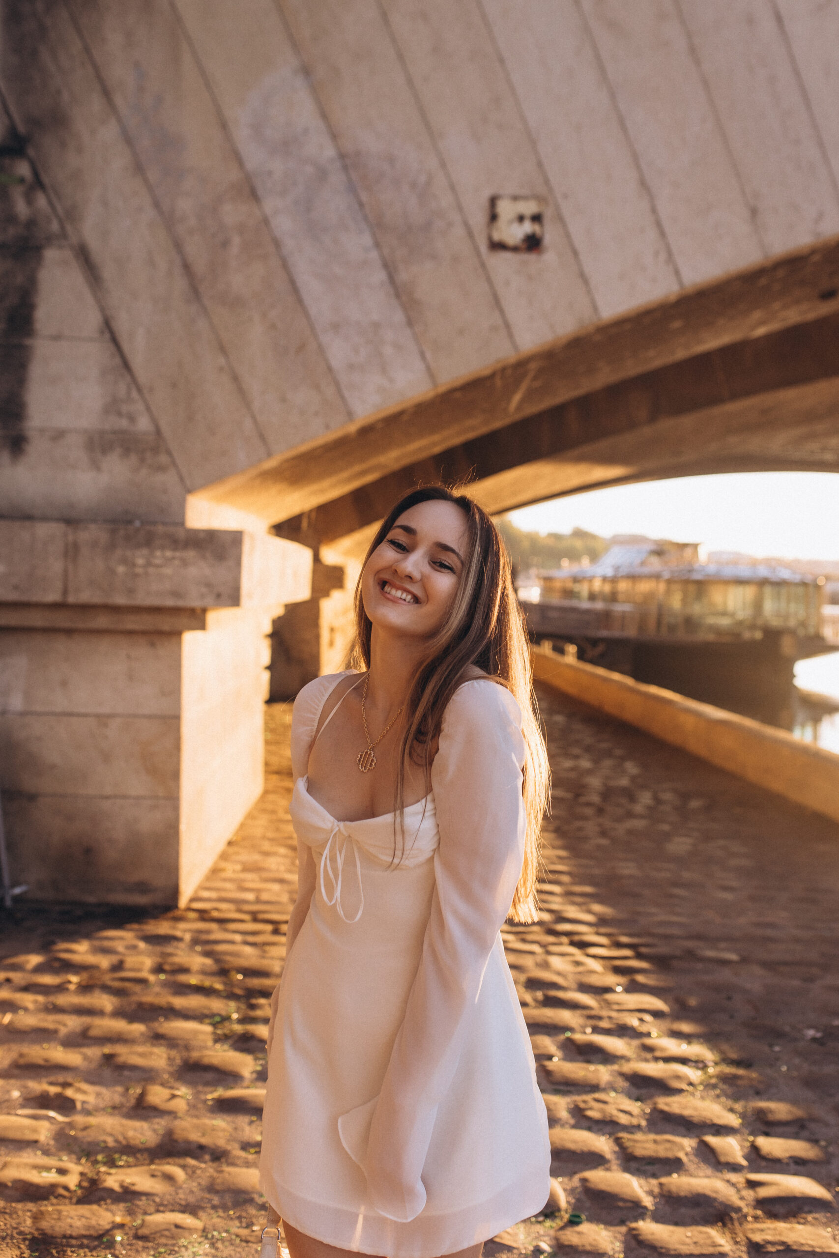 A candid solo photoshoot beneath a Parisian bridge arch, golden hour light glowing across old cobblestones by the Seine. Photographed by Julia Litvin, litvinphoto.com.