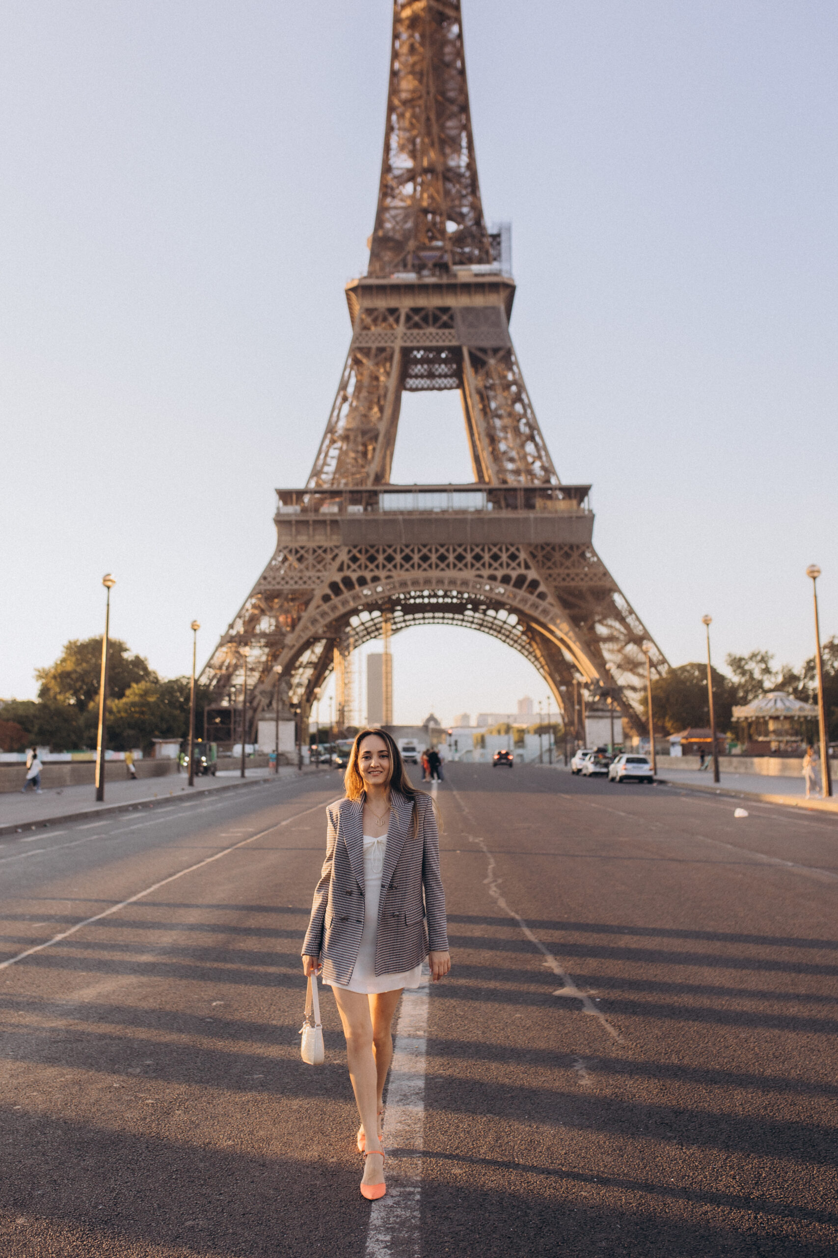 A woman in a white dress and checked blazer walking down an empty avenue straight toward the Eiffel Tower.