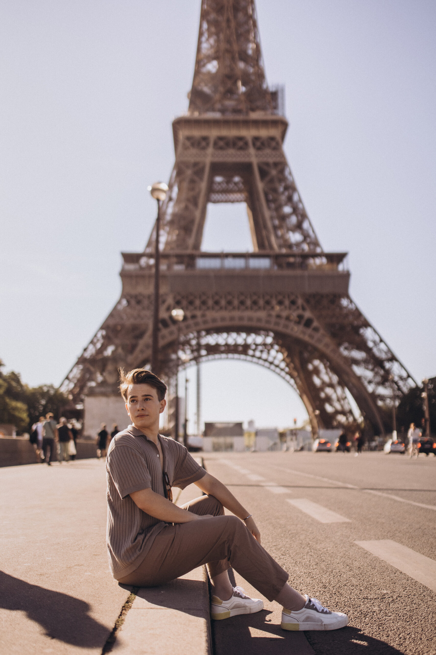 A young man sitting on a curb on the open esplanade, the full Eiffel Tower rising behind him in bright daylight.