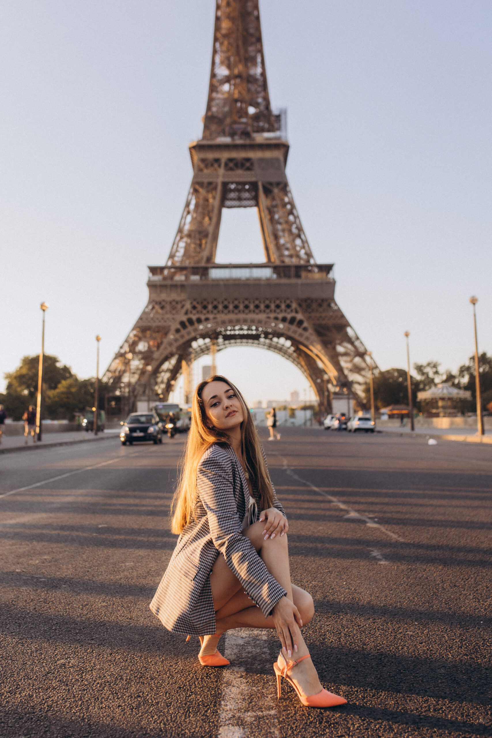 A woman in a checked blazer crouching on an empty avenue at golden hour, the Eiffel Tower filling the frame behind her.