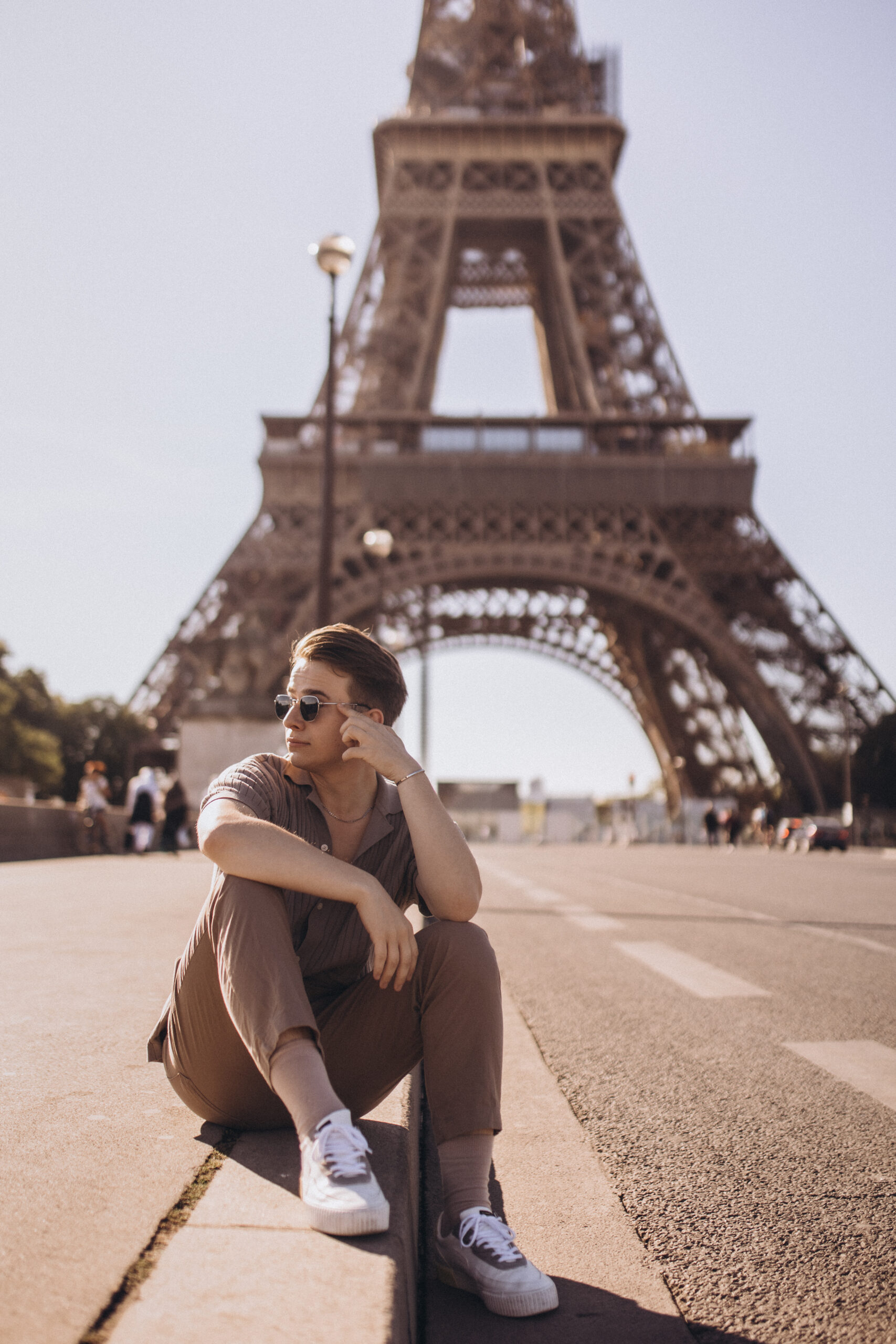 A men's lifestyle photoshoot at the foot of the Eiffel Tower, a young man seated on the esplanade in bright daylight with the landmark close behind. Photographed by Julia Litvin, litvinphoto.com.