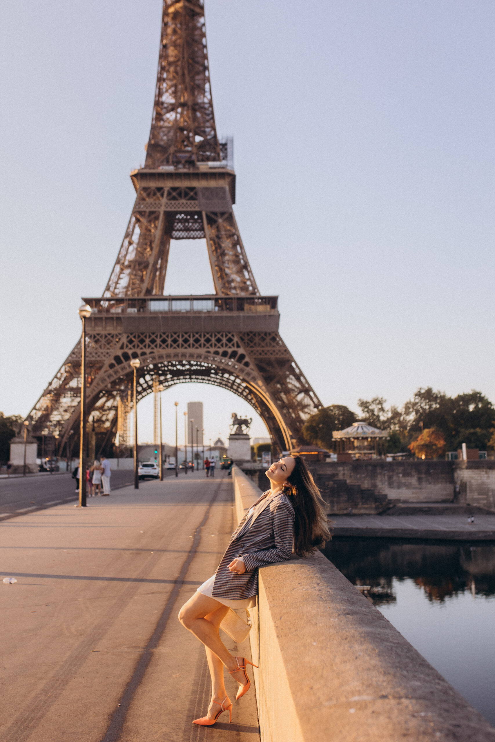 A woman in a checked blazer leaning back against a bridge parapet, the Eiffel Tower rising through its arch behind her.