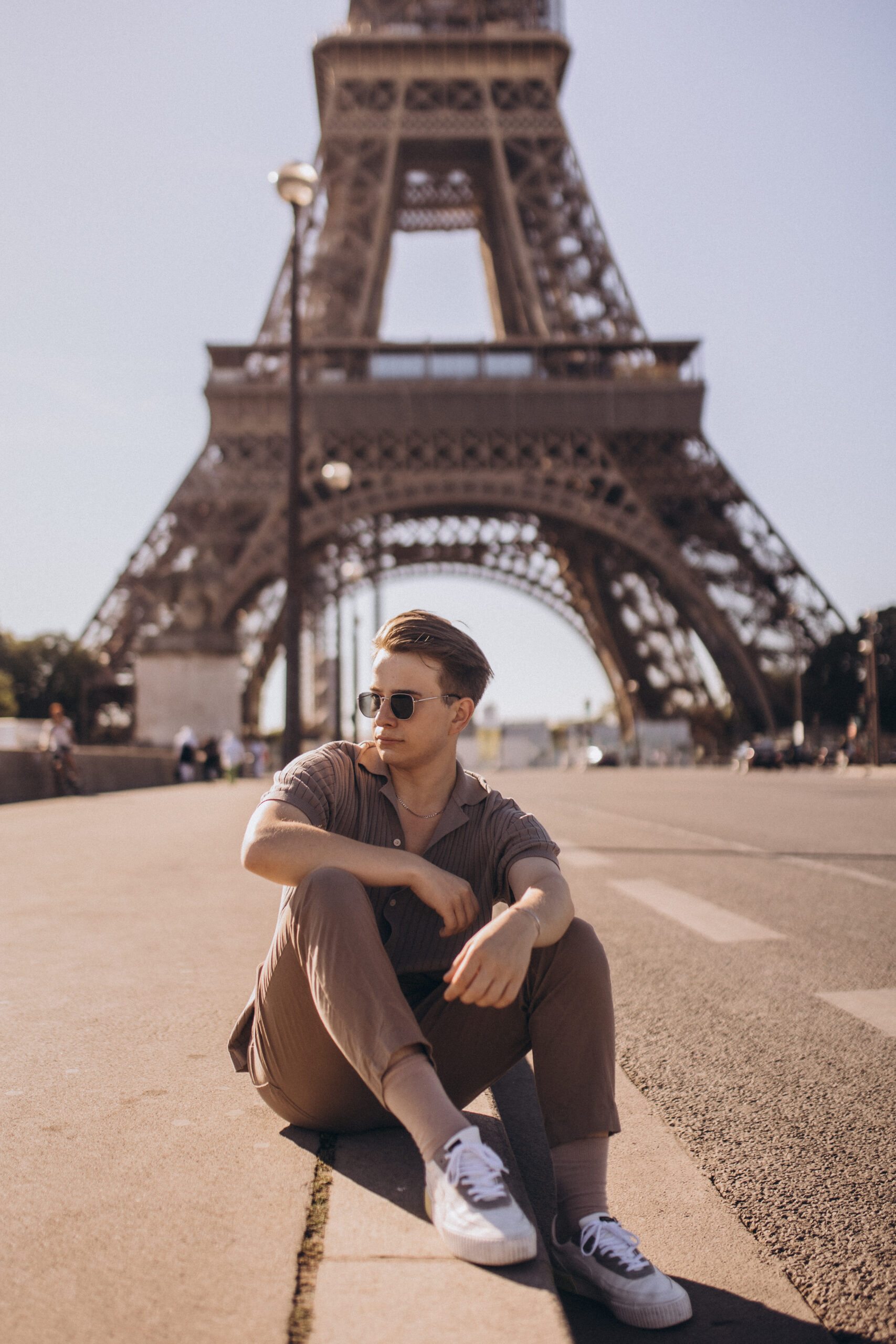 A young man in sunglasses sitting on the esplanade in front of the Eiffel Tower, resting an arm on his knee in bright daylight.