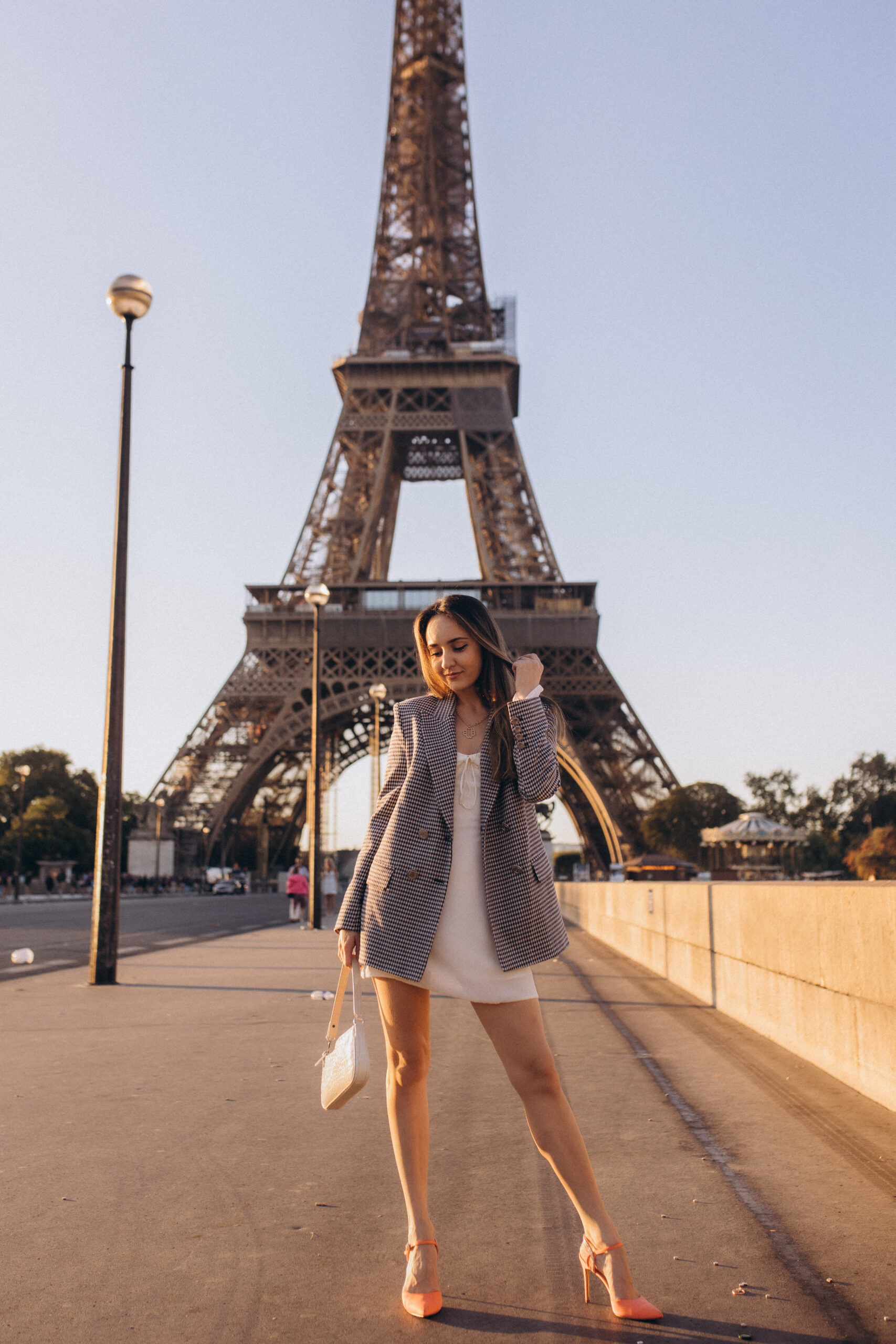 A stylish full-length photoshoot on a Paris bridge, a checked blazer layered over a white dress with the Eiffel Tower centered behind. Photographed by Julia Litvin, litvinphoto.com.