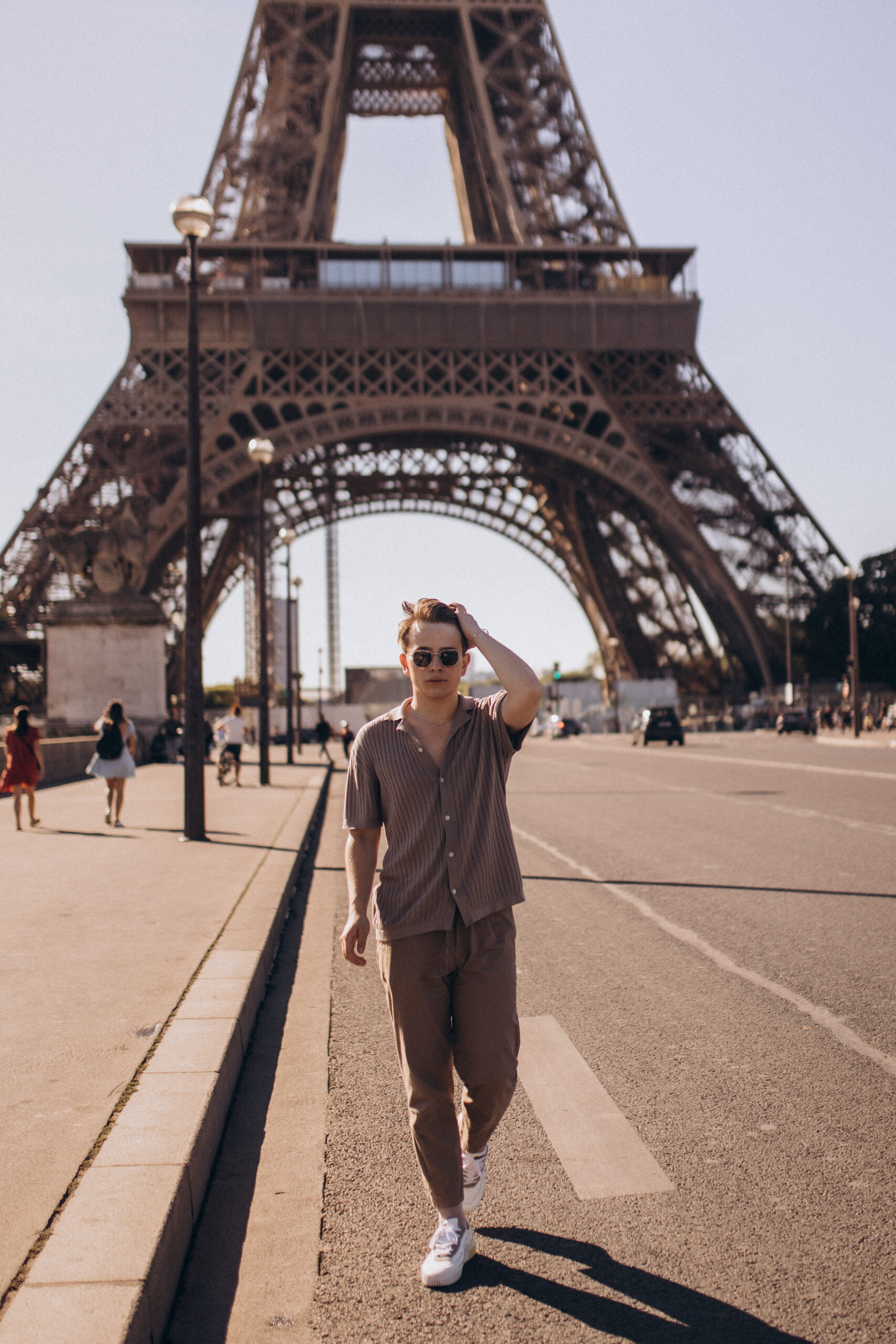 A young man walking across the open esplanade toward the camera, running a hand through his hair with the Eiffel Tower behind him.