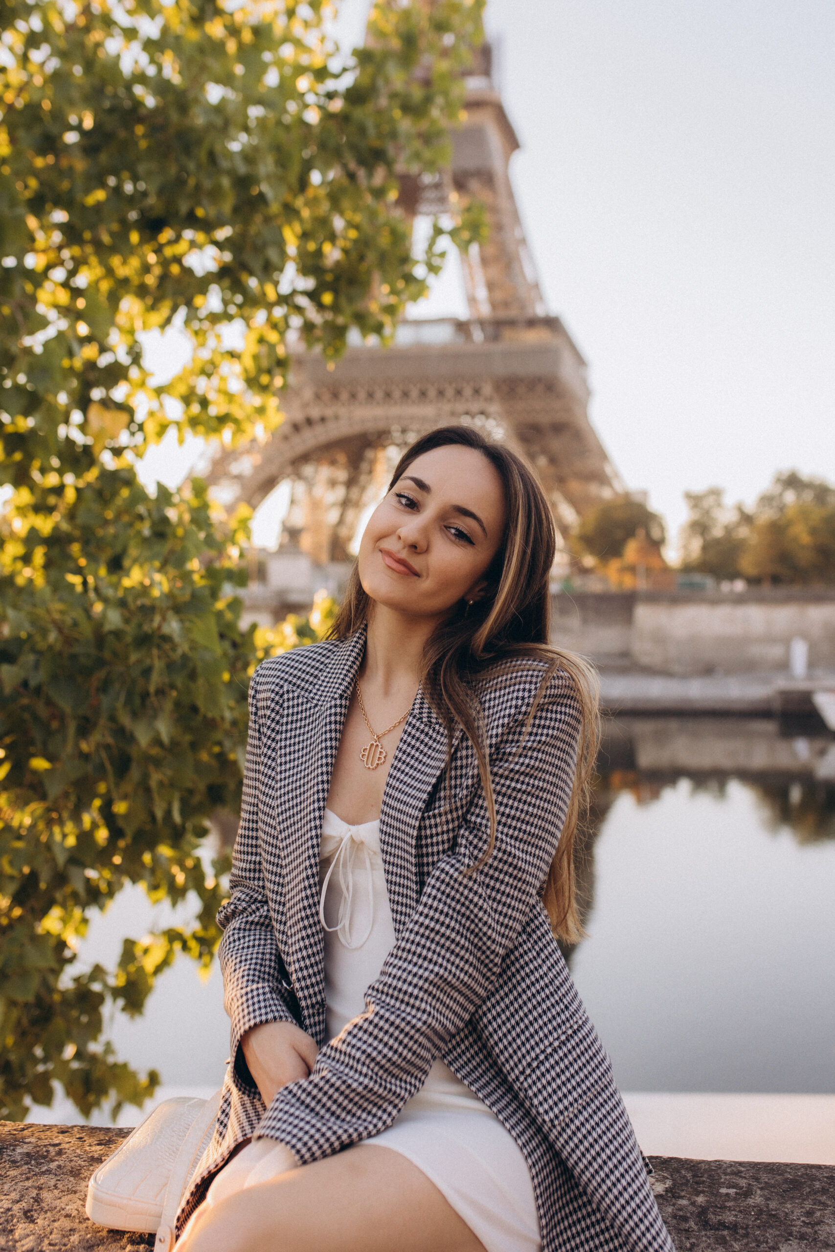 A tender individual portrait by the Seine, a checked blazer and white dress framed by turning leaves with the Eiffel Tower beyond. Photographed by Julia Litvin, litvinphoto.com.