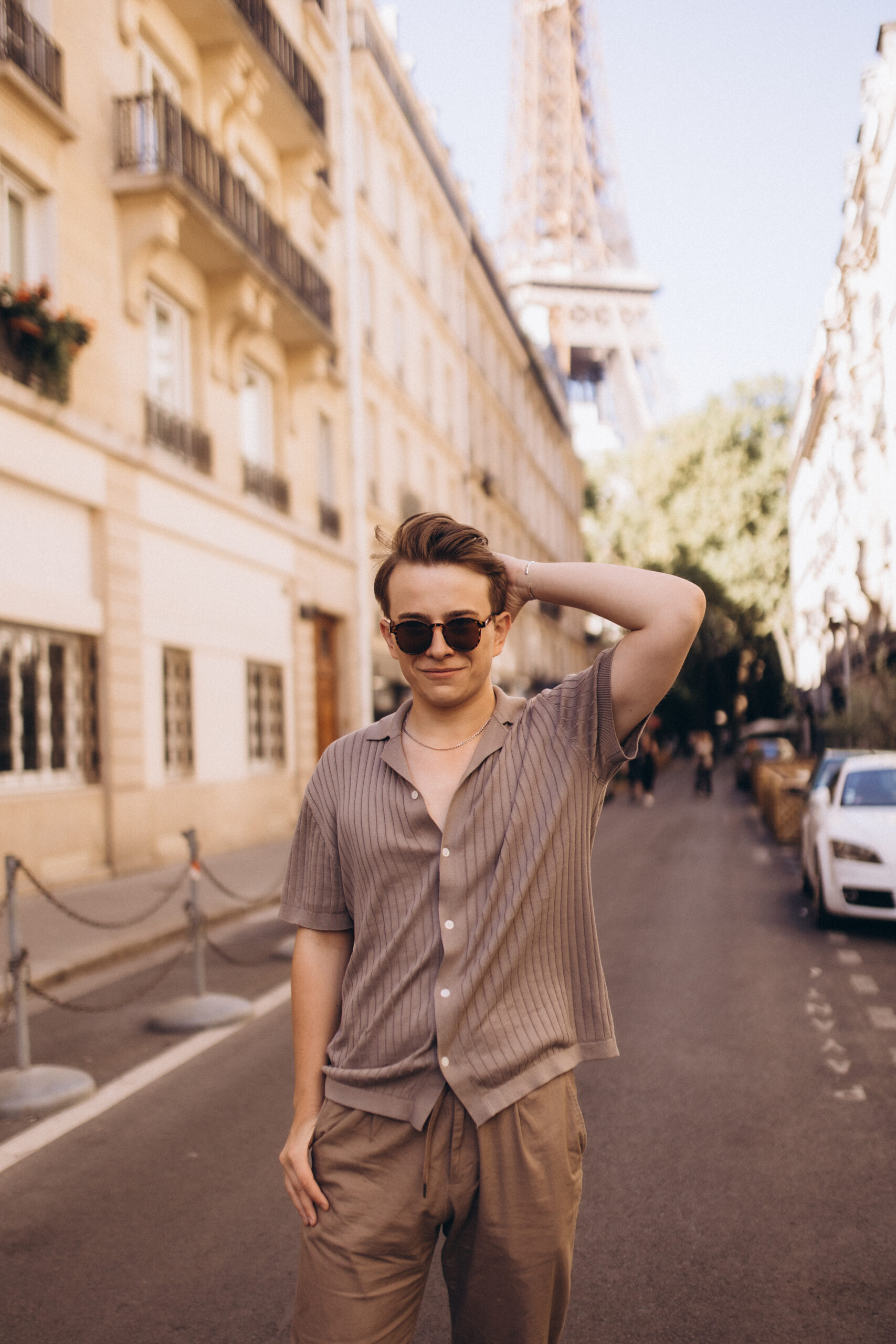 A relaxed male street-style portrait in Paris, a young man in sunglasses and a beige knit shirt with the Eiffel Tower in the background. Photographed by Julia Litvin, litvinphoto.com.