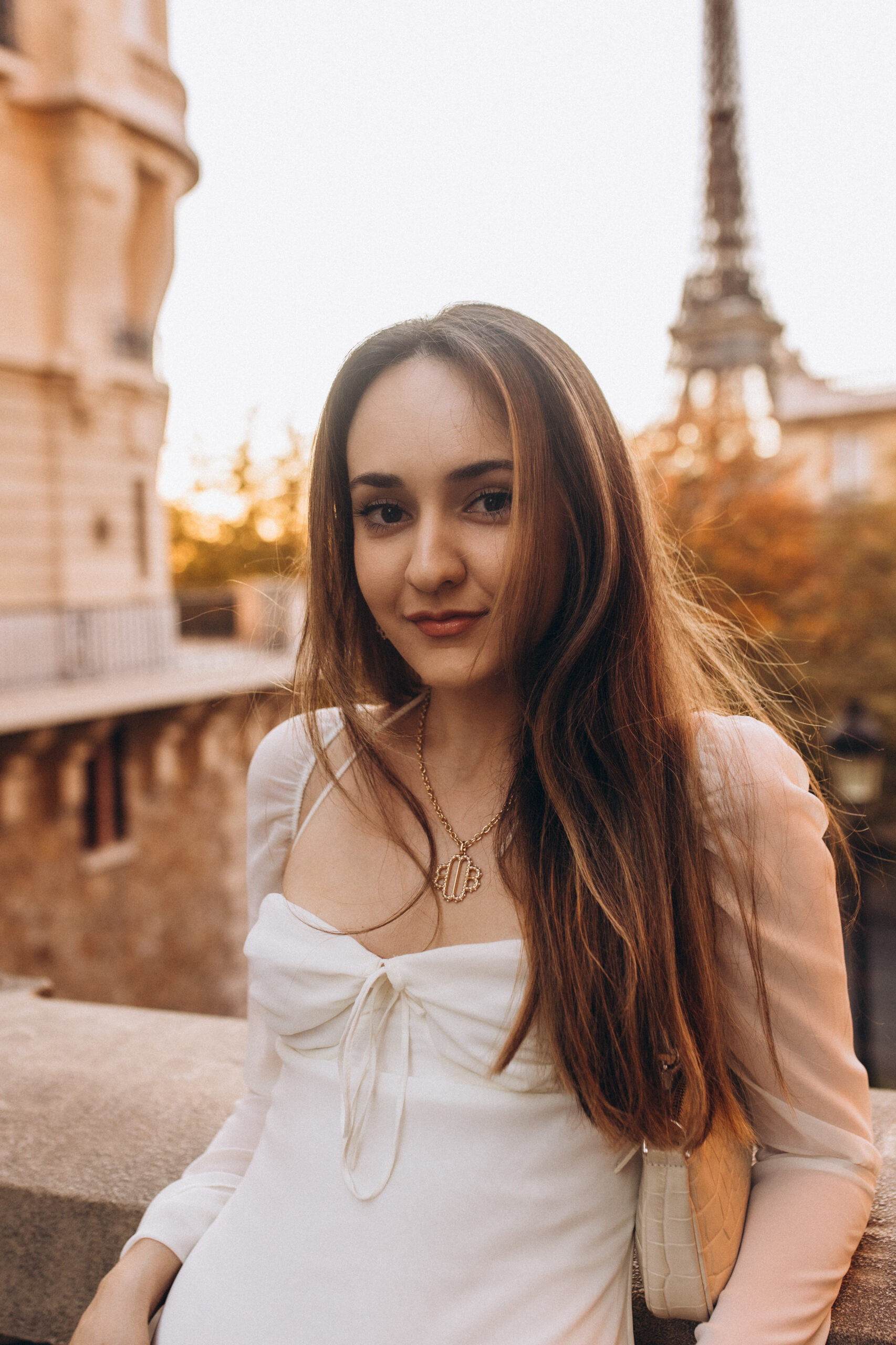 A woman in a white dress smiling gently at golden hour, the Eiffel Tower glowing in the warm light behind her.