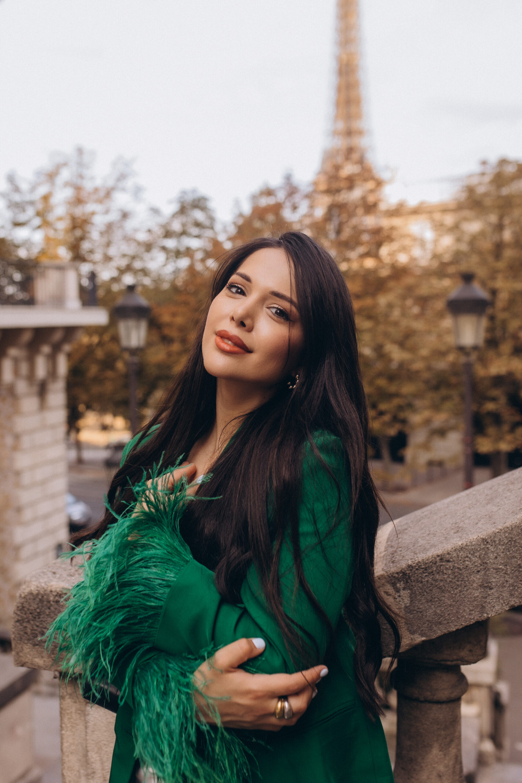 Portrait photoshoot in Paris, a woman in an emerald feathered blazer leaning on a stone balustrade, gazing at the camera, autumn foliage behind her