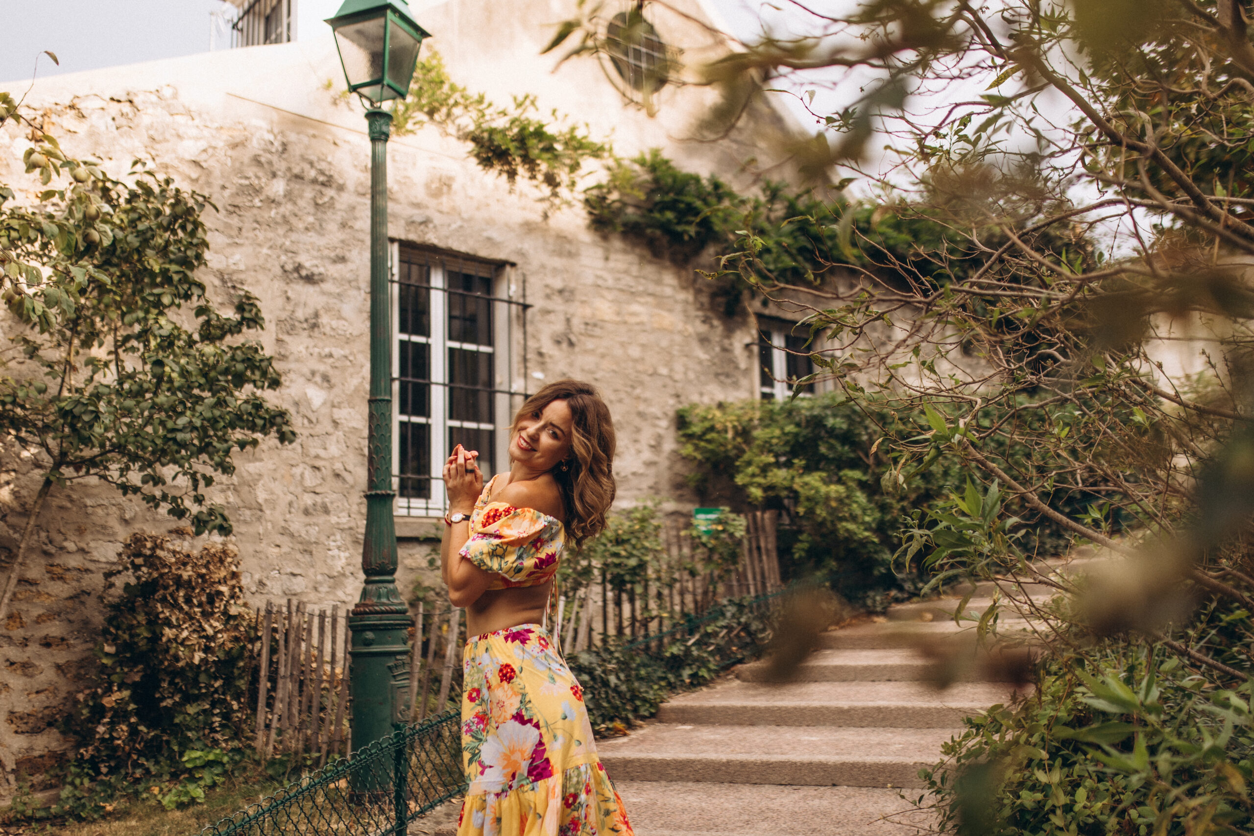 Montmartre photoshoot in Paris, a woman in a yellow floral dress glancing back over her shoulder on a green, leafy Montmartre lane