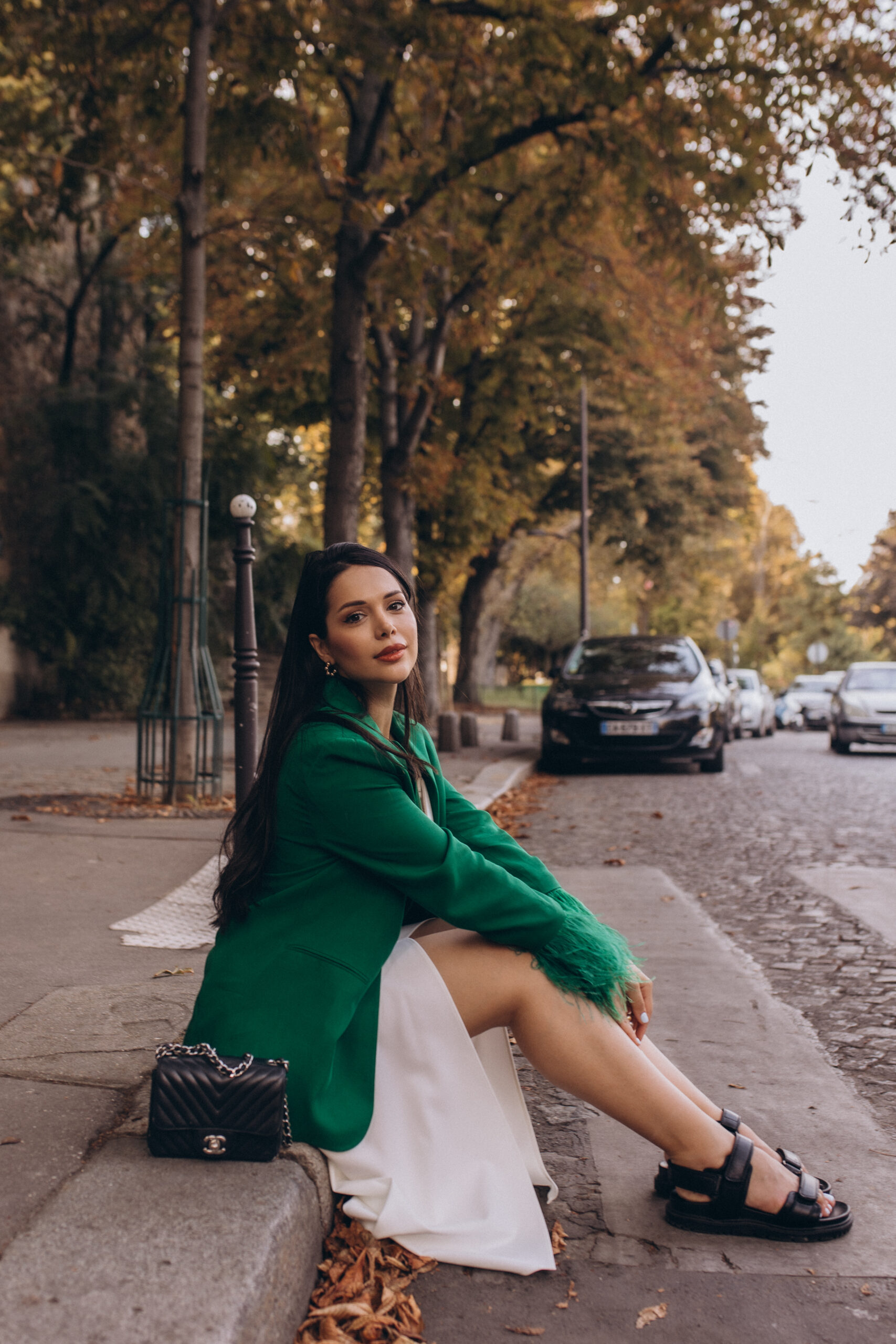 Paris portrait session, a woman in a green feathered blazer and white dress sitting on a curb of a tree-lined Parisian street in autumn