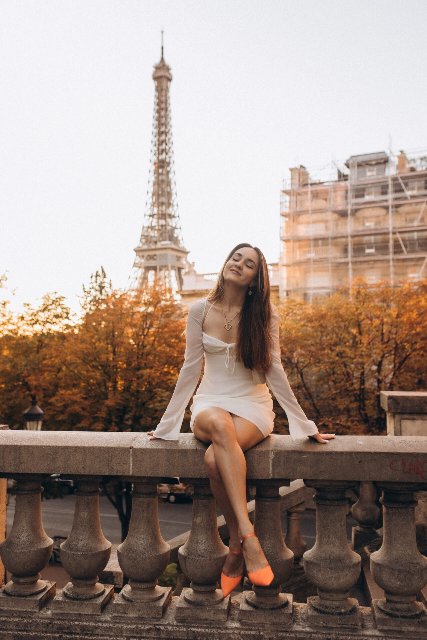 A peaceful solo photoshoot in Paris at golden hour, a woman in white seated on a balustrade among autumn foliage with the Eiffel Tower behind. Photographed by Julia Litvin, litvinphoto.com