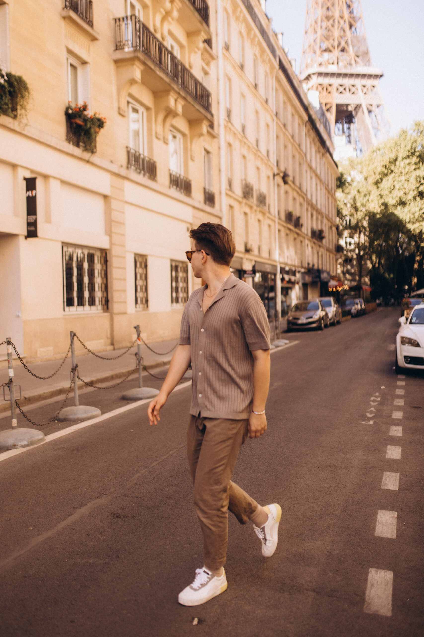 A young man in a beige shirt and chinos walking across a sunlit Parisian street, the Eiffel Tower behind the rooftops.