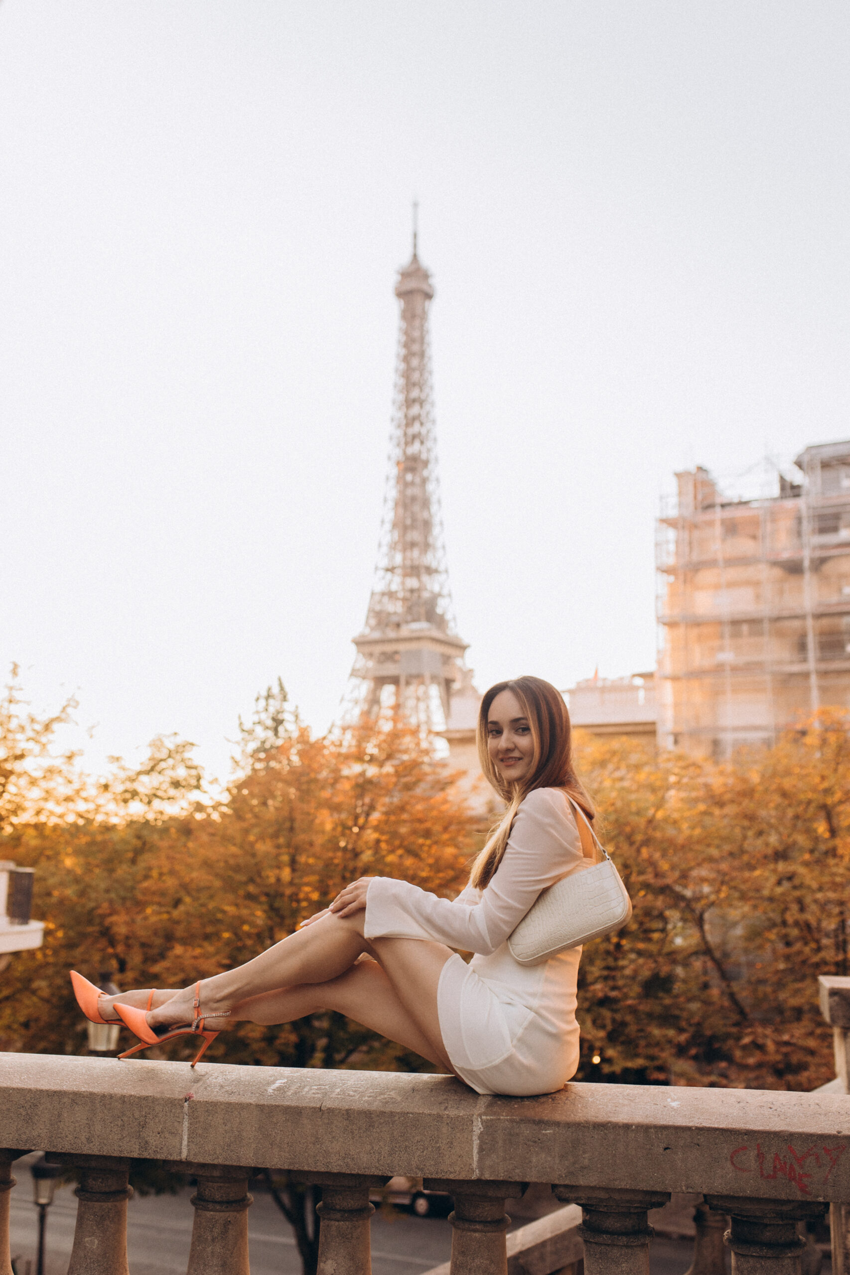A woman in a white dress and orange heels sitting on a stone balustrade, autumn trees and the Eiffel Tower behind her.