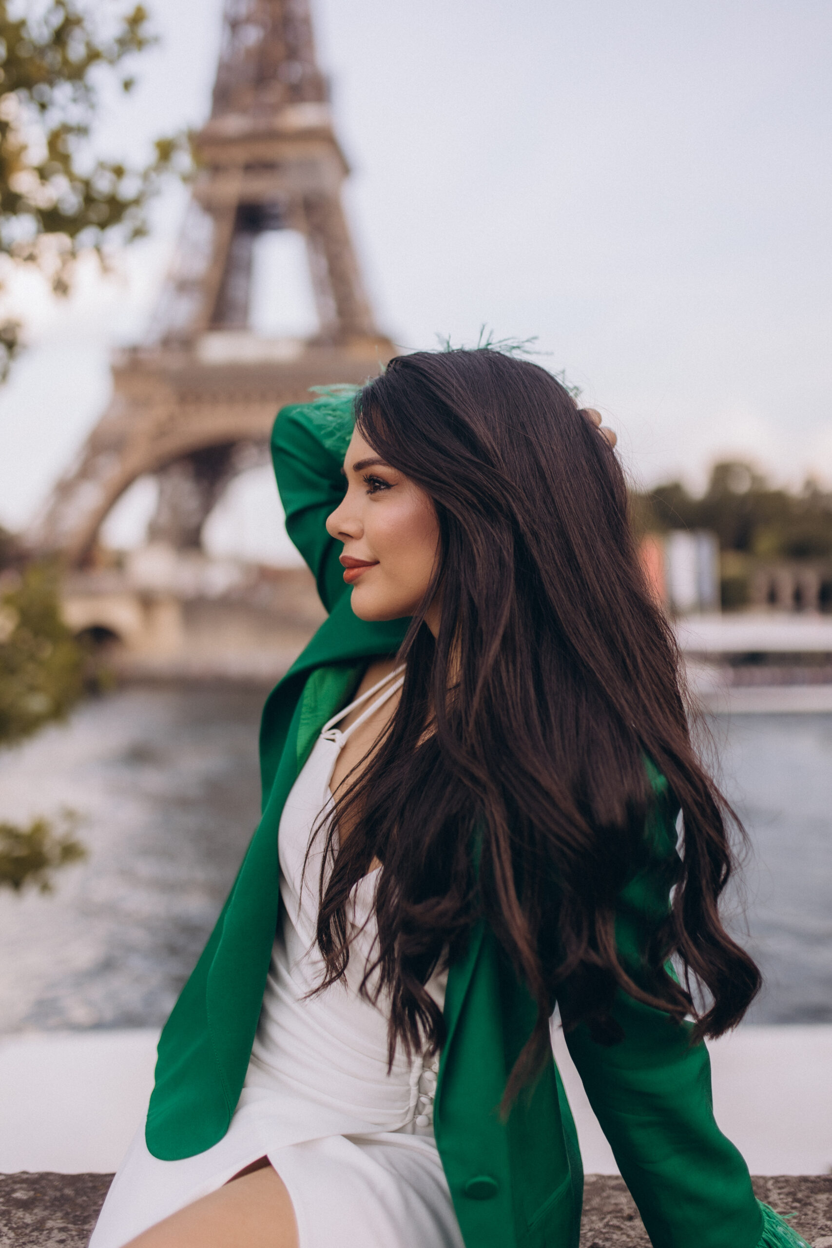 Portrait photoshoot in Paris by the Seine, a woman in a green blazer and white slip dress in profile, a famous iron landmark rising across the river