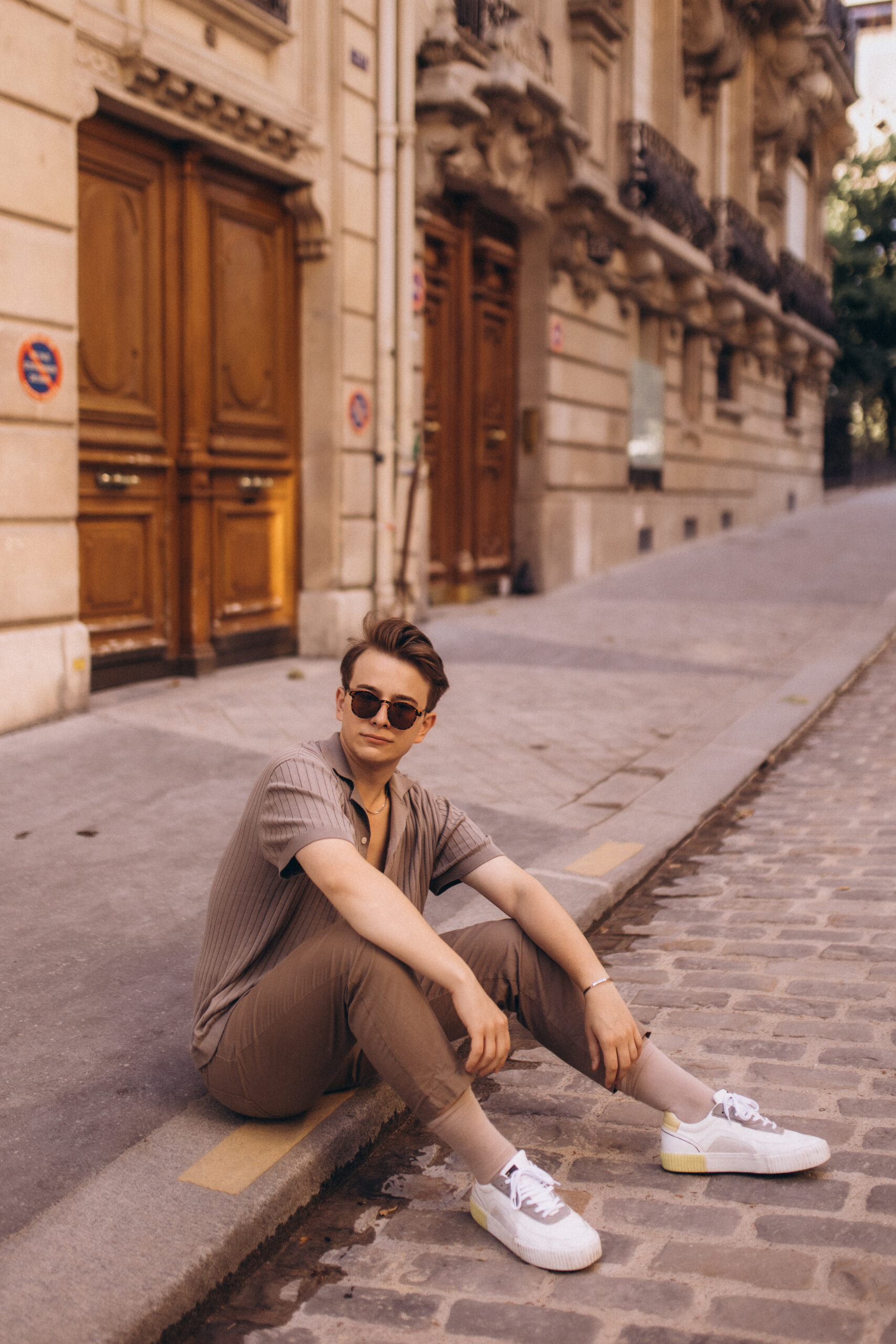 Street photography in France, a young man in sunglasses sitting on the edge of a cobblestone Paris street in front of tall carved wooden doors