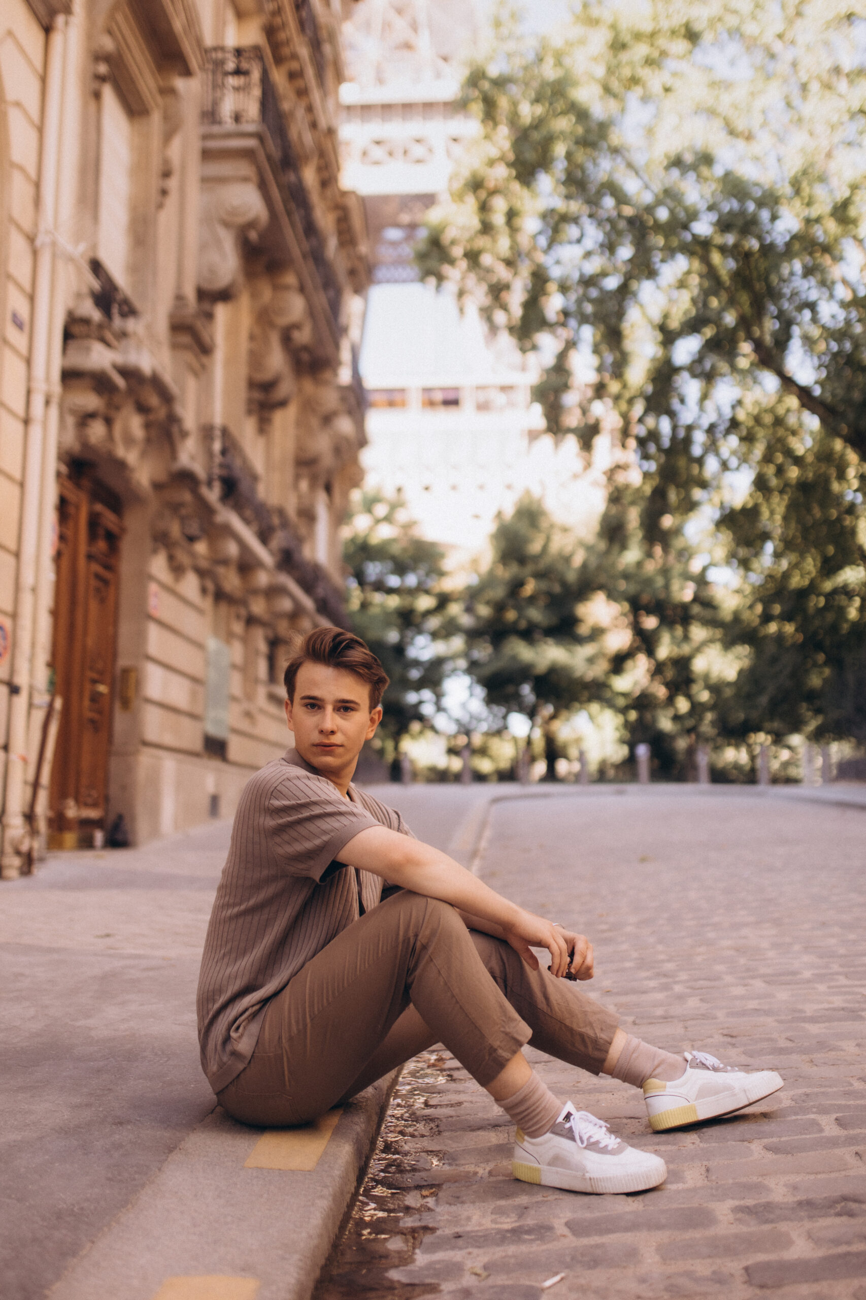 A young man sitting on a cobblestone curb on a Paris street, the base of the Eiffel Tower framed by green trees behind him.