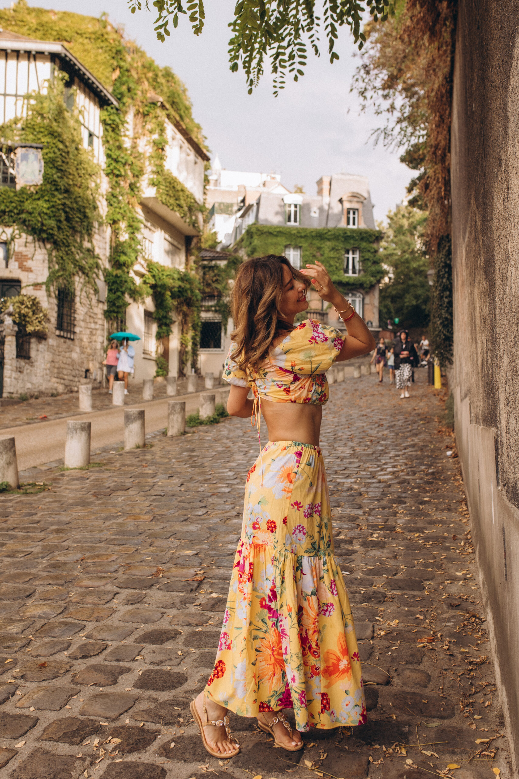 Montmartre photoshoot in Paris on Rue de l'Abreuvoir, a woman in a yellow floral two-piece walking away down the ivy-lined cobblestone lane, one hand lifted to her hair