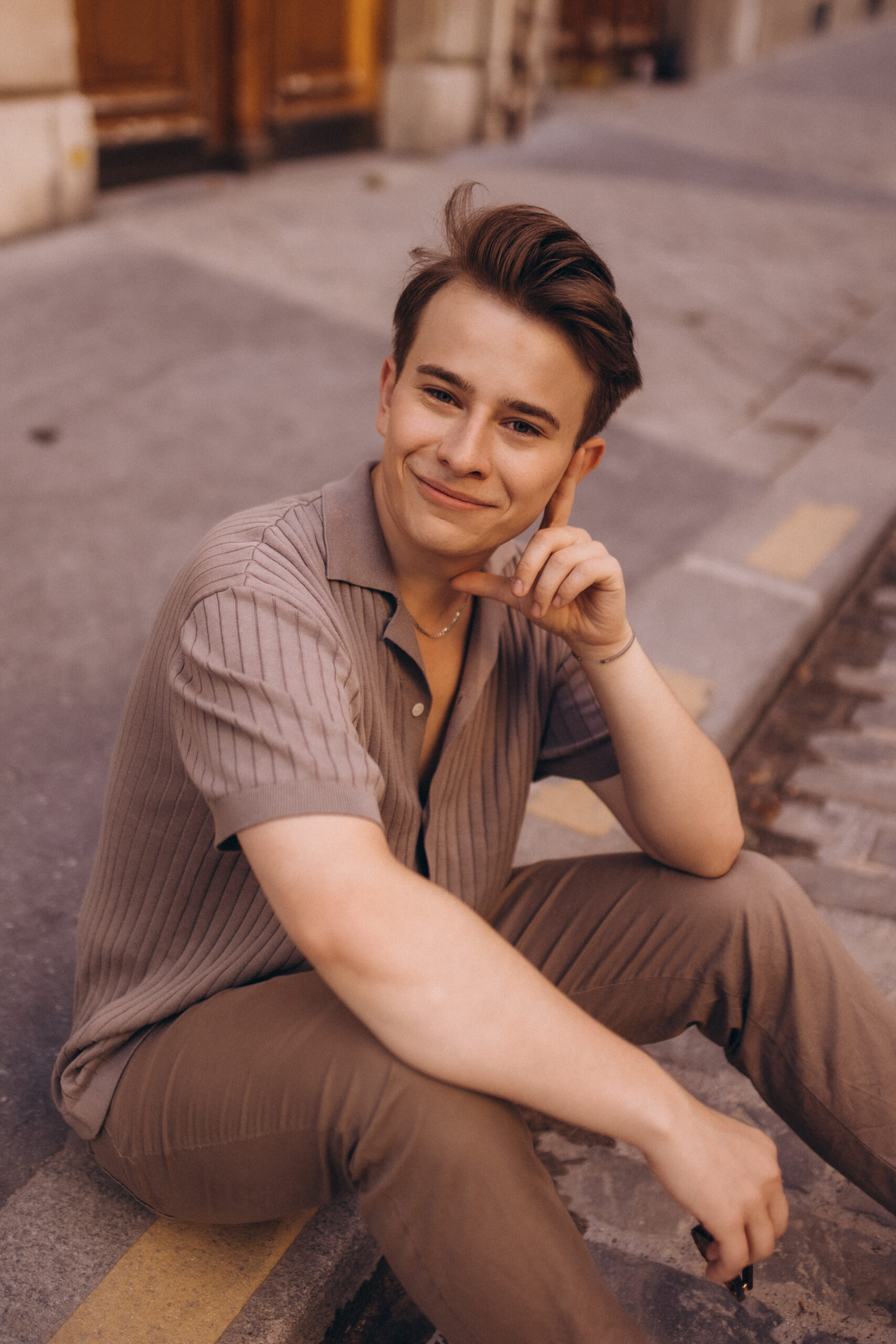 A close-up portrait of a smiling young man in a beige knit shirt sitting on a Parisian street, chin resting on his hand.