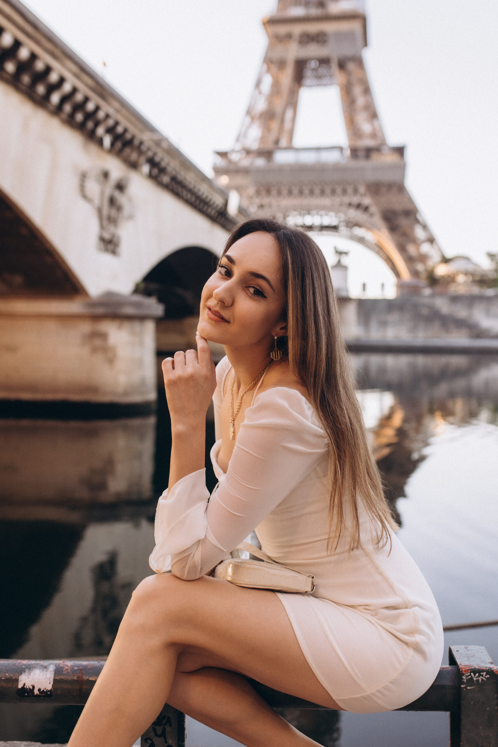 A serene individual portrait by the Seine in Paris, seated near a stone arch bridge with the Eiffel Tower rising close behind. Photographed by Julia Litvin, litvinphoto.com.
