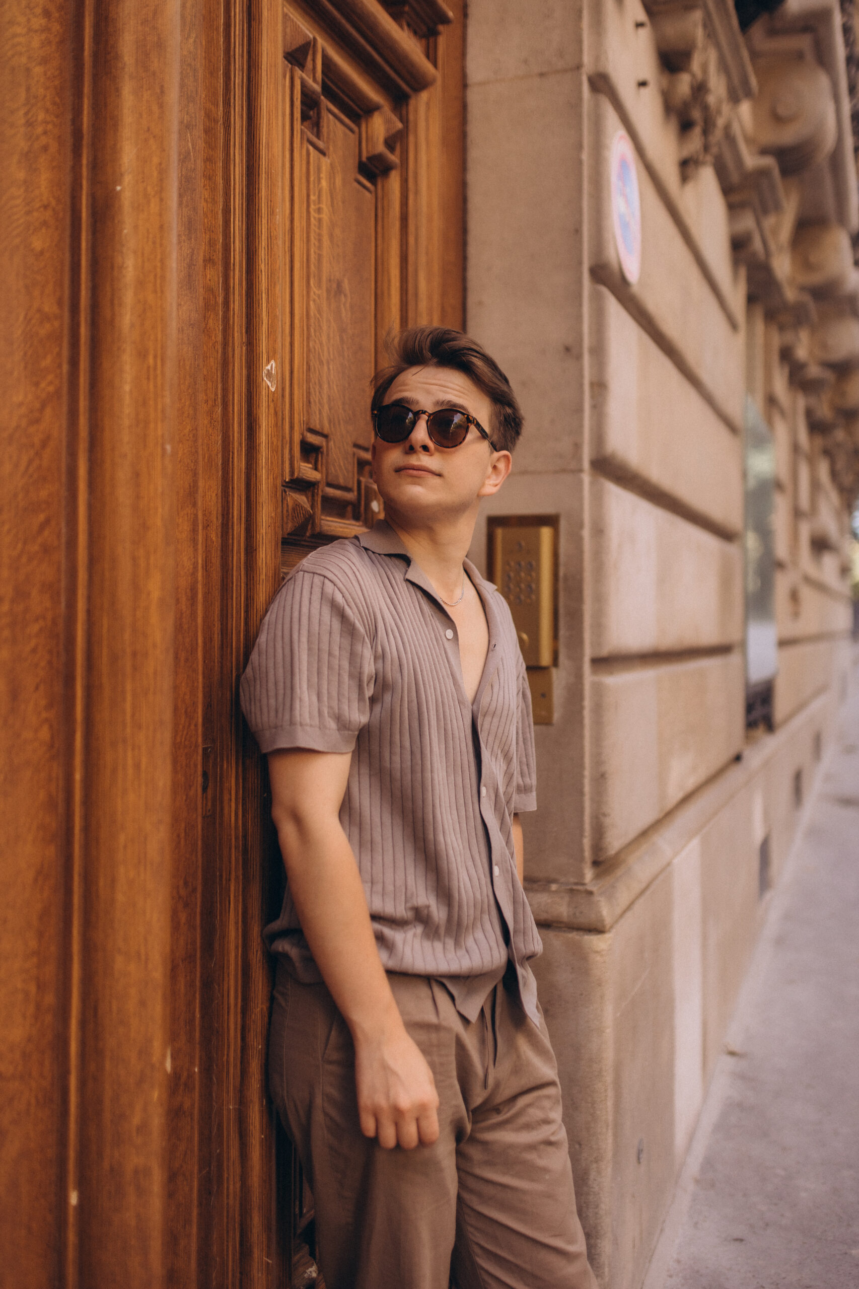 A young man in sunglasses leaning against a tall carved wooden door on a Paris street, gazing upward.